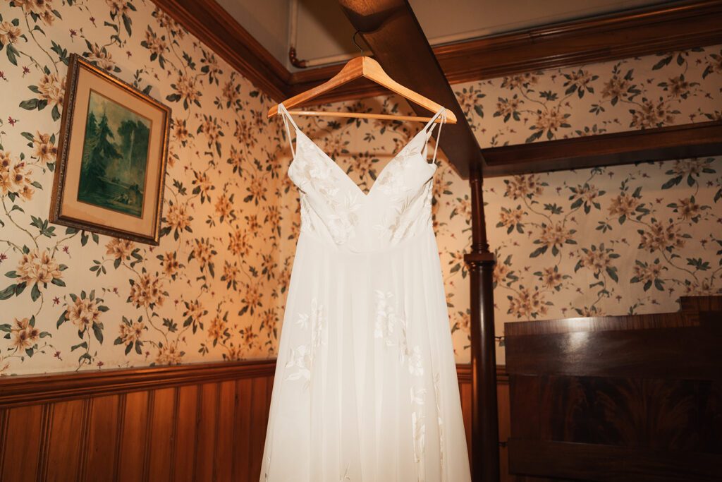 a wedding dress hangs on the bed frame among a wooden room with delicate floral wallpaper at newlywed couple intimating leans into each other among rose gardens at Belhurst Castle and Winery