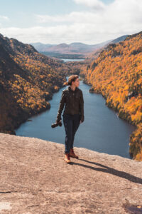 a female photographer holding her camera overlooking autumn mountains and a river in the Adirondack Mountains of new york