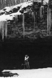 a newly engaged couple cuddles for warmth in each others arms under a frozen waterfall in round top new york