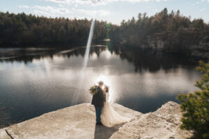 a newlywed couple hugs each other on a cliff side overlooking Minnewaska State Park Preserve lake on their elopement day. the couple wears western inspired wedding outfits featuring boho cowboy hats and boots.