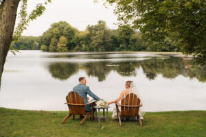 a bride and groom sit on adirondack chairs on their wedding day overlooking a lake