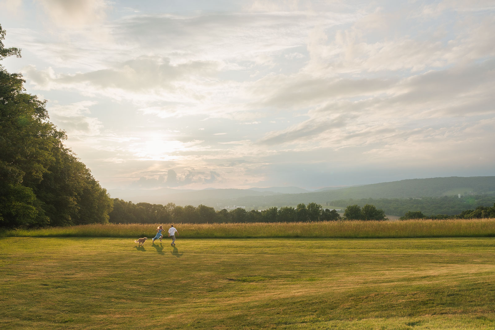 a couple runs around in a wide open field looking over mountains in new jersey with their dog after a sun shower